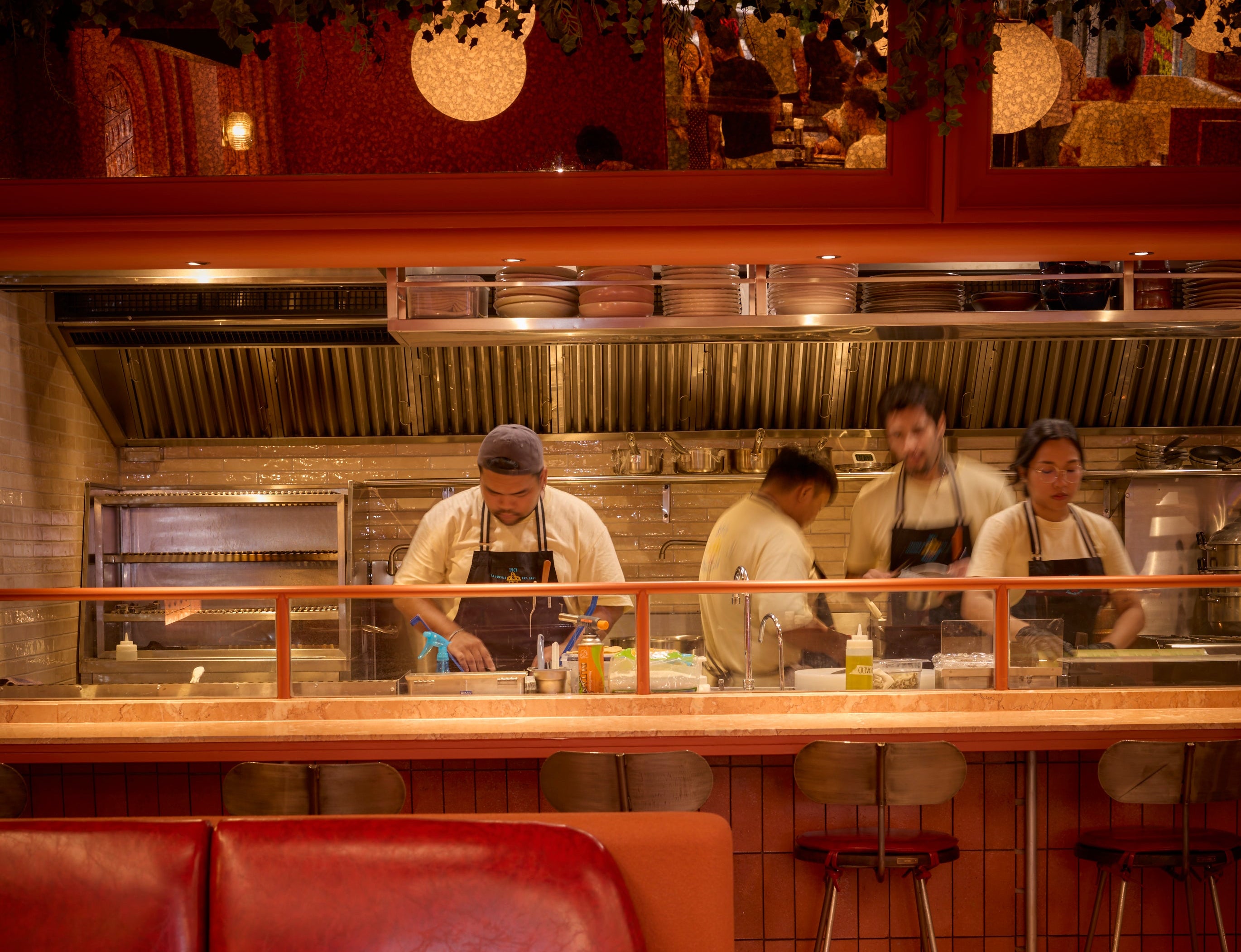 A warm, atmospheric restaurant interior with red leather booths in the foreground, a wooden counter, and three chefs working behind a glass partition in a kitchen area.